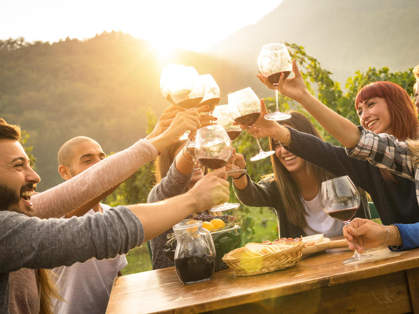 Happy friends having fun outdoors - Young people enjoying harvest time together at farmhouse vineyard countryside - Youth and friendship concept - Focus on hands toasting wine glasses with sun flare